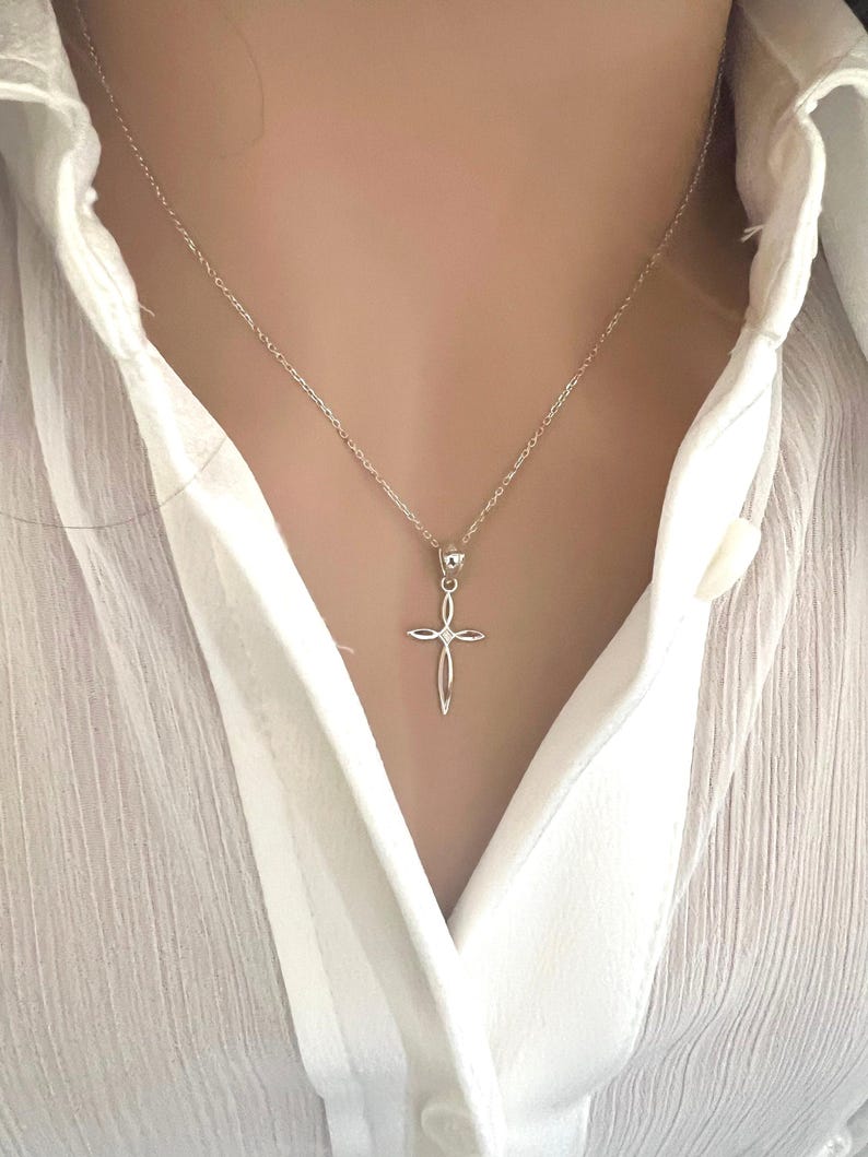 Puede incluir: Un collar con colgante de cruz plateada. La cruz tiene un dise&ntilde;o calado con un peque&ntilde;o acento redondo en el centro. El collar se lleva sobre una camisa blanca.