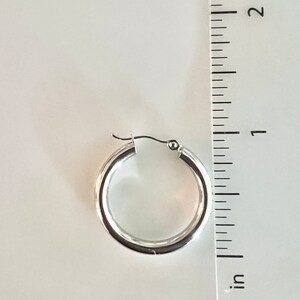 K&ouml;nnte beinhalten: Ein einzelner silberner Creolen-Ohrring mit einem Durchmesser von etwa 2,5 cm wird vor einem wei&szlig;en Hintergrund gezeigt. Der Ohrring hat eine polierte, reflektierende Oberfl&auml;che und ein schlichtes, klassisches Design. Ein Lineal liegt zur Gr&ouml;&szlig;enbestimmung neben dem Ohrring.