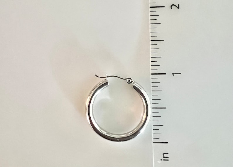 K&ouml;nnte beinhalten: Ein einzelner silberner Creolen-Ohrring mit einem Durchmesser von etwa 2,5 cm wird vor einem wei&szlig;en Hintergrund gezeigt. Der Ohrring hat eine polierte, reflektierende Oberfl&auml;che und ein schlichtes, klassisches Design. Ein Lineal liegt zur Gr&ouml;&szlig;enbestimmung neben dem Ohrring.