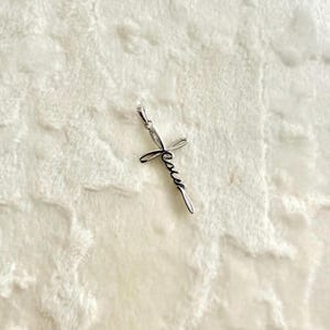 May include: A silver-colored pendant in the shape of a cross. The word "Jesus" is written in cursive script across the cross. The pendant has a small loop at the top for a chain.