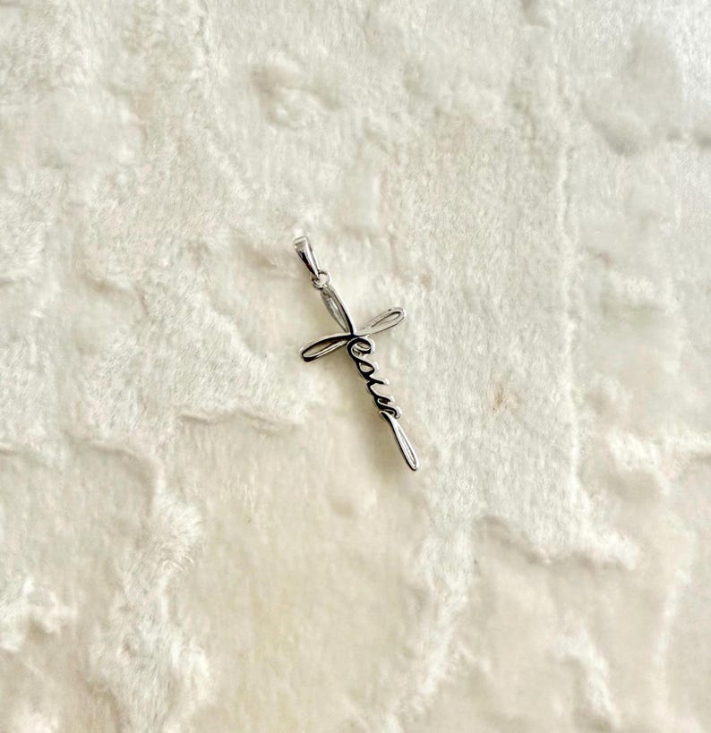 May include: A silver-colored pendant in the shape of a cross. The word "Jesus" is written in cursive script across the cross. The pendant has a small loop at the top for a chain.