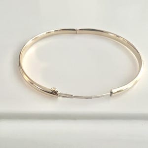 May include: A gold-colored hinged bangle bracelet. The bracelet is round and has a simple, elegant design. The clasp is visible and the bracelet is on a white surface.