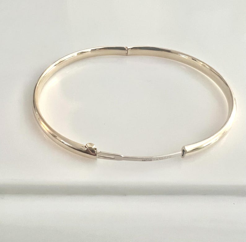 May include: A gold-colored hinged bangle bracelet. The bracelet is round and has a simple, elegant design. The clasp is visible and the bracelet is on a white surface.