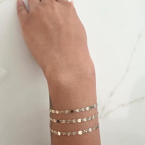 May include: Three delicate gold bracelets adorn a wrist. Each bracelet features a repeating pattern of small, flat, oval and star-shaped links. The bracelets are layered, creating a subtle, elegant accessory. The wrist is set against a white marble surface.