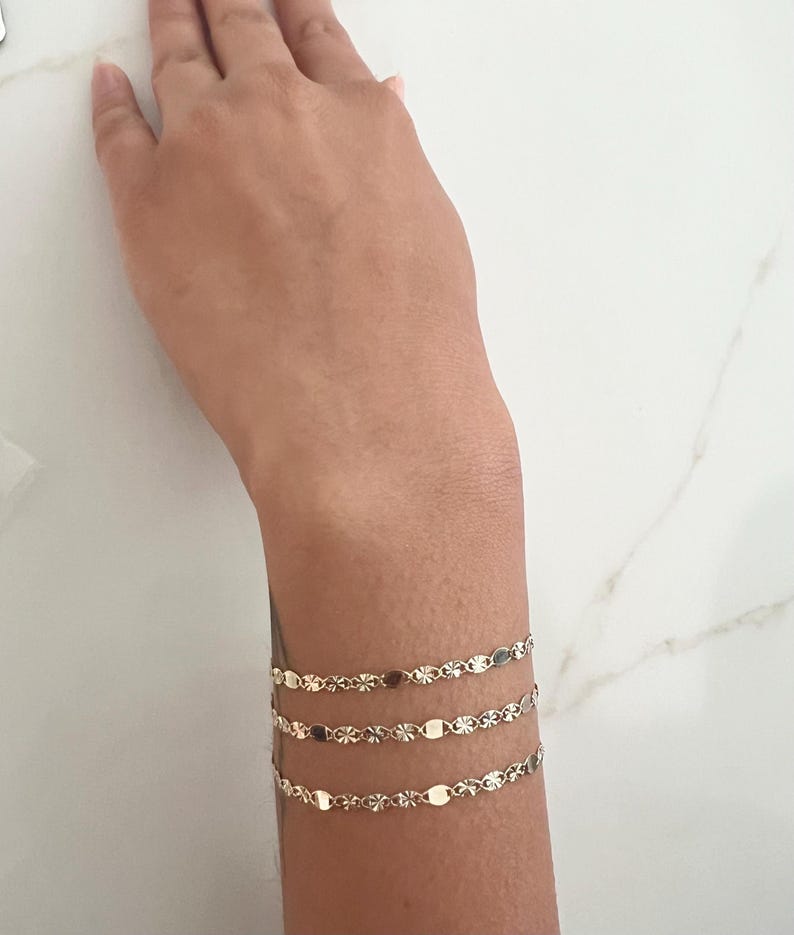 May include: Three delicate gold bracelets adorn a wrist. Each bracelet features a repeating pattern of small, flat, oval and star-shaped links. The bracelets are layered, creating a subtle, elegant accessory. The wrist is set against a white marble surface.