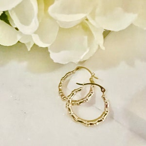 Puede incluir: Un par de pendientes de aro dorados con un dise&ntilde;o similar al bamb&uacute;. Los pendientes est&aacute;n colocados sobre una superficie de m&aacute;rmol blanco con un fondo borroso de flores blancas.