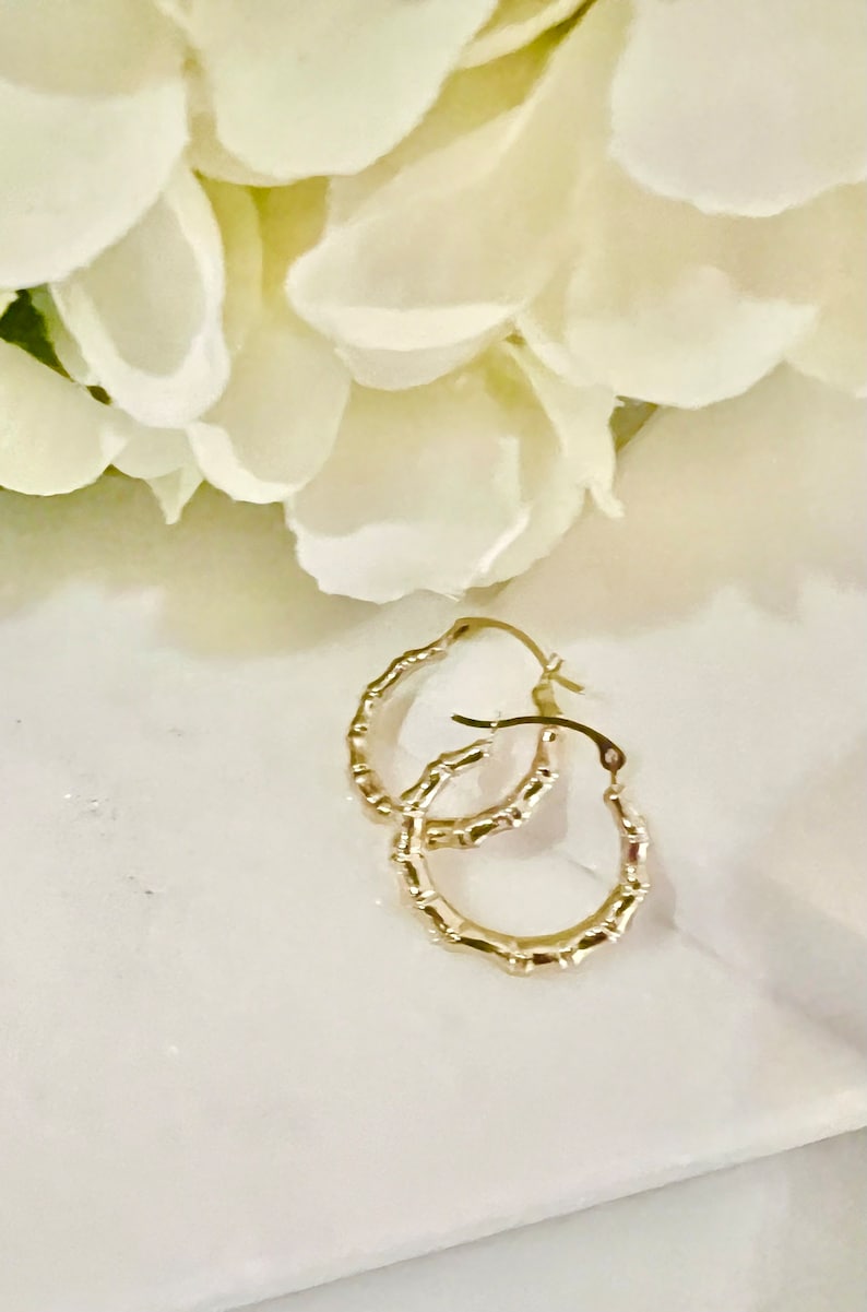 Puede incluir: Un par de pendientes de aro dorados con un dise&ntilde;o similar al bamb&uacute;. Los pendientes est&aacute;n colocados sobre una superficie de m&aacute;rmol blanco con un fondo borroso de flores blancas.