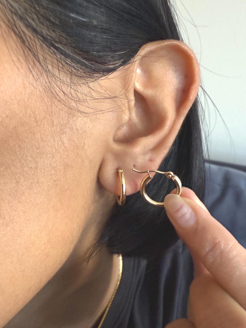 Op de afbeelding: Een oor met een kleine gouden oorring. Een andere gouden oorring wordt tegen het oor gehouden. De oorbellen zijn eenvoudig en klassiek van ontwerp, met een gepolijste afwerking. De persoon draagt een donkere top.