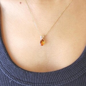 May include: A gold necklace with a teardrop-shaped, orange-colored pendant. The pendant is suspended from a delicate gold chain. The necklace is worn against a dark blue ribbed tank top.