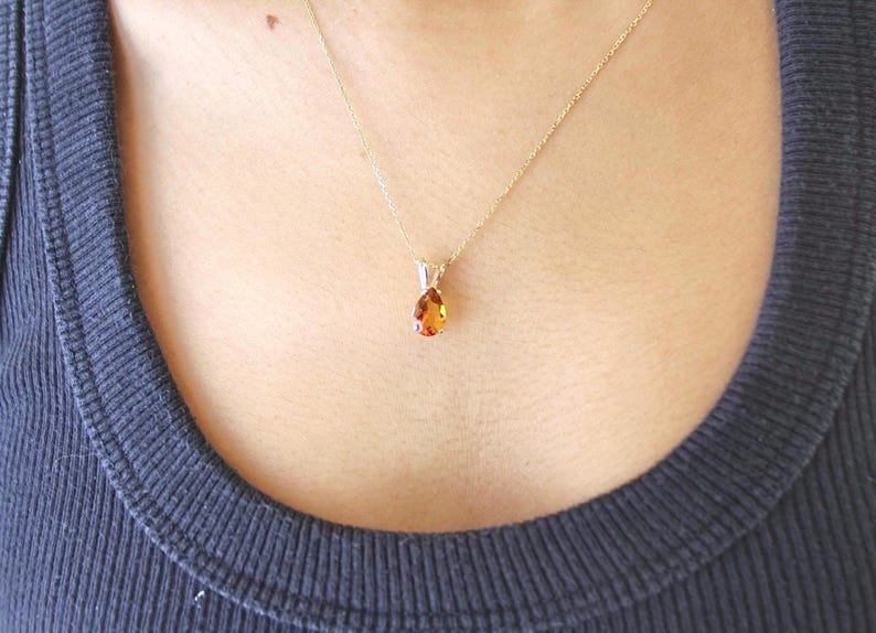 May include: A gold necklace with a teardrop-shaped, orange-colored pendant. The pendant is suspended from a delicate gold chain. The necklace is worn against a dark blue ribbed tank top.