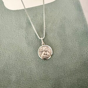 Peut inclure: Un collier pendentif argent&eacute; avec un m&eacute;daillon rond repr&eacute;sentant un ange. L'ange, avec des ailes, repose sa t&ecirc;te sur sa main. La cha&icirc;ne est une fine cha&icirc;ne argent&eacute;e.