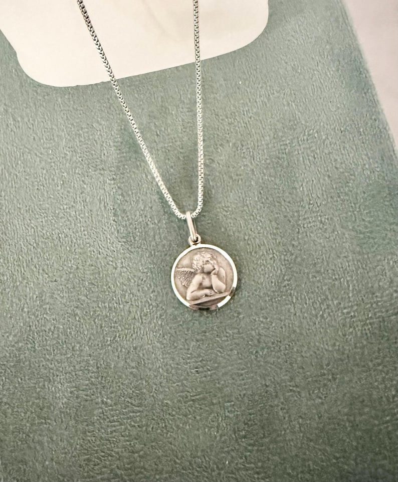 Peut inclure: Un collier pendentif argent&eacute; avec un m&eacute;daillon rond repr&eacute;sentant un ange. L'ange, avec des ailes, repose sa t&ecirc;te sur sa main. La cha&icirc;ne est une fine cha&icirc;ne argent&eacute;e.