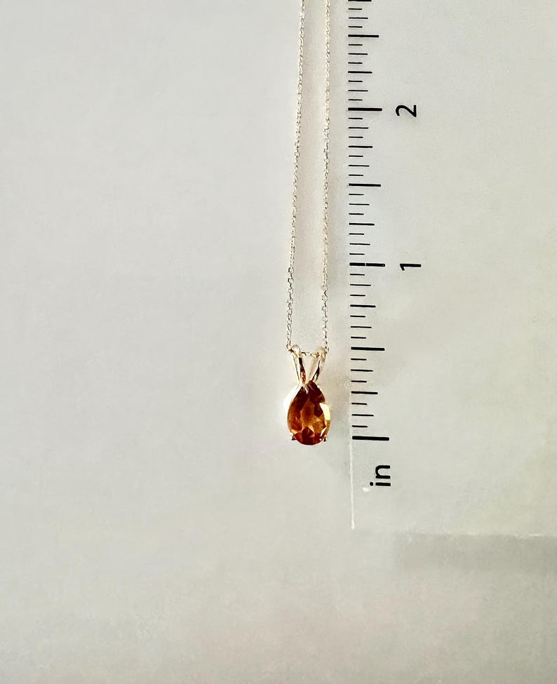 May include: A gold-colored necklace with a teardrop-shaped, amber-colored gemstone pendant. The pendant is approximately 0.5 inches in length. The necklace is displayed against a white background.