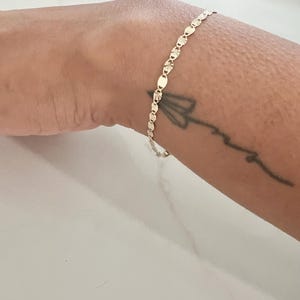 May include: A delicate gold bracelet featuring a series of small, textured oval links. The bracelet is worn on a wrist with a small tattoo. The bracelet's design is simple and elegant, suitable for everyday wear.