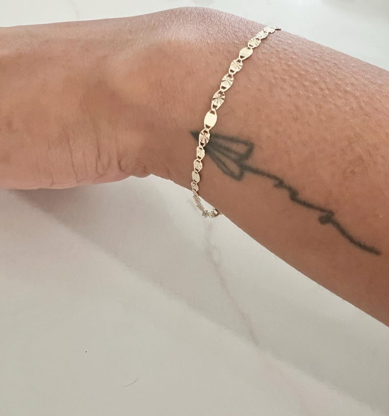 May include: A delicate gold bracelet featuring a series of small, textured oval links. The bracelet is worn on a wrist with a small tattoo. The bracelet's design is simple and elegant, suitable for everyday wear.