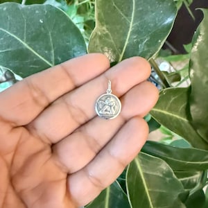 Peut inclure: Un petit pendentif rond en argent repr&eacute;sentant un ch&eacute;rubin en relief. Le ch&eacute;rubin repose sa t&ecirc;te sur sa main, avec des ailes visibles. Le pendentif est tenu dans une main, sur un fond de feuilles vertes.