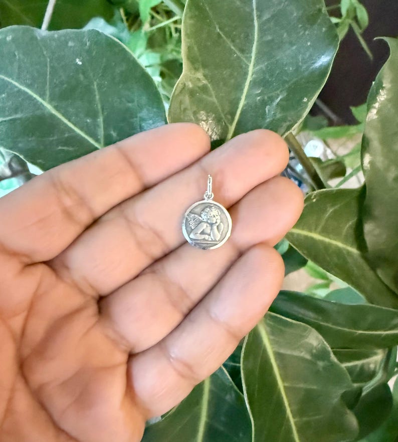 Peut inclure: Un petit pendentif rond en argent repr&eacute;sentant un ch&eacute;rubin en relief. Le ch&eacute;rubin repose sa t&ecirc;te sur sa main, avec des ailes visibles. Le pendentif est tenu dans une main, sur un fond de feuilles vertes.