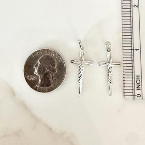 May include: Two silver-colored cross pendants with the word "Jesus" written vertically. A quarter coin and an inch ruler are next to the pendants. The pendants are approximately 1 inch (2.5 cm) in length.