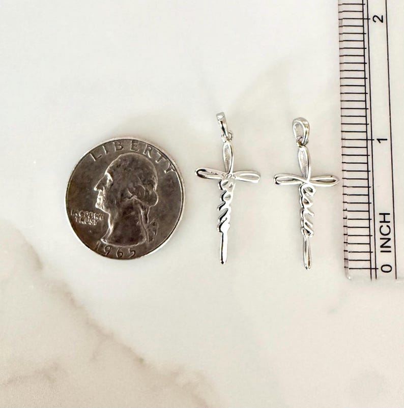May include: Two silver-colored cross pendants with the word "Jesus" written vertically. A quarter coin and an inch ruler are next to the pendants. The pendants are approximately 1 inch (2.5 cm) in length.