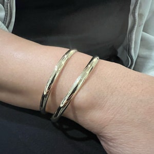 May include: Two gold-colored, open-ended bangles are worn on a wrist. The bangles have a smooth, polished surface and a rounded shape. The jewelry is simple and elegant, reflecting light.