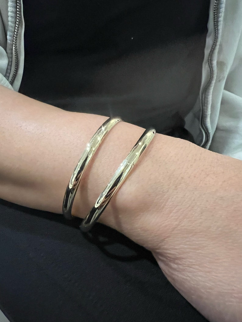 May include: Two gold-colored, open-ended bangles are worn on a wrist. The bangles have a smooth, polished surface and a rounded shape. The jewelry is simple and elegant, reflecting light.