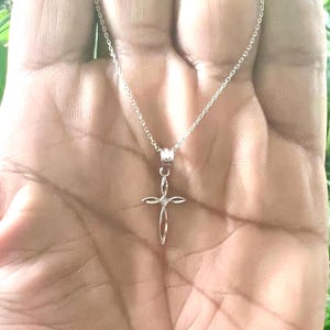 Puede incluir: Un collar plateado con un delicado colgante de cruz. El dise&ntilde;o de la cruz es abierto y delgado, con una peque&ntilde;a gema en el centro. El collar se muestra sobre un fondo de tono de piel.