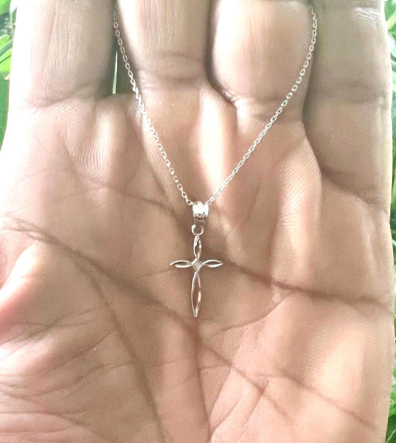 Puede incluir: Un collar plateado con un delicado colgante de cruz. El dise&ntilde;o de la cruz es abierto y delgado, con una peque&ntilde;a gema en el centro. El collar se muestra sobre un fondo de tono de piel.
