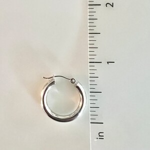 K&ouml;nnte beinhalten: Ein einzelner silberner Creolen-Ohrring. Der Ohrring hat einen Durchmesser von ca. 2,5 cm. Der Ohrring hat ein schlichtes, poliertes Design und einen Klappverschluss. Der Hintergrund ist hell und neutral.