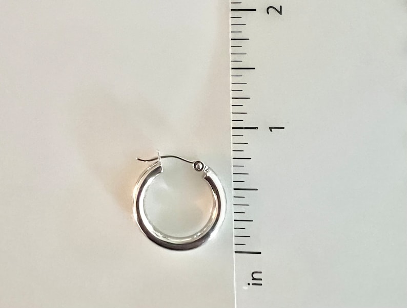 K&ouml;nnte beinhalten: Ein einzelner silberner Creolen-Ohrring. Der Ohrring hat einen Durchmesser von ca. 2,5 cm. Der Ohrring hat ein schlichtes, poliertes Design und einen Klappverschluss. Der Hintergrund ist hell und neutral.
