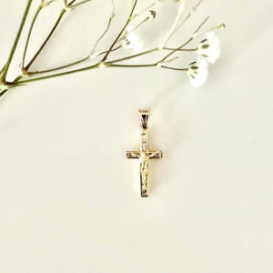 Op de afbeelding: Een goudkleurige kruisvormige hanger met een kleine afbeelding van Jezus Christus. Het kruis is versierd met kleine, heldere edelstenen. Delicate witte bloemen en stelen bevinden zich linksboven op een zachte, gebroken witte achtergrond.