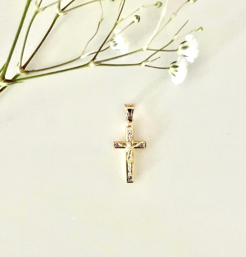 Op de afbeelding: Een goudkleurige kruisvormige hanger met een kleine afbeelding van Jezus Christus. Het kruis is versierd met kleine, heldere edelstenen. Delicate witte bloemen en stelen bevinden zich linksboven op een zachte, gebroken witte achtergrond.