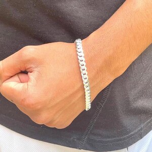 May include: A silver curb chain bracelet. The bracelet is made of interlocking links and is worn on a wrist. The background is a black shirt.