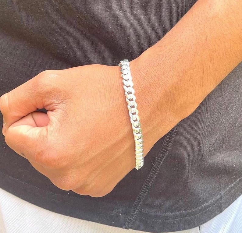 May include: A silver curb chain bracelet. The bracelet is made of interlocking links and is worn on a wrist. The background is a black shirt.