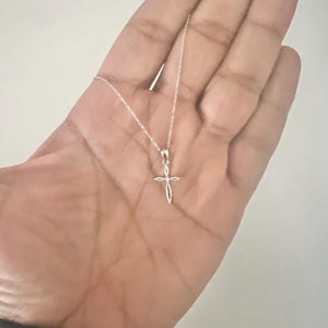 Puede incluir: Un delicado collar con colgante de cruz de plata. La cruz presenta un dise&ntilde;o abierto y minimalista suspendido de una fina cadena. El collar se muestra sobre un fondo neutro, destacando la simple elegancia de la joya.
