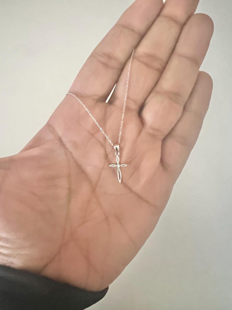 Puede incluir: Un delicado collar con colgante de cruz de plata. La cruz presenta un dise&ntilde;o abierto y minimalista suspendido de una fina cadena. El collar se muestra sobre un fondo neutro, destacando la simple elegancia de la joya.