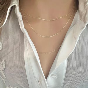 May include: Three gold chain necklaces layered on a person's neck. The chains are delicate and have a subtle shimmer.