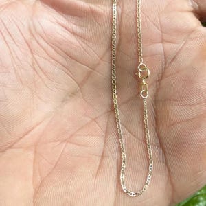 May include: A delicate gold chain bracelet featuring small, oval-shaped mariner links. The bracelet is secured with a circular spring ring clasp and includes a small rectangular metal tag near the closure, showcasing a polished metallic finish.