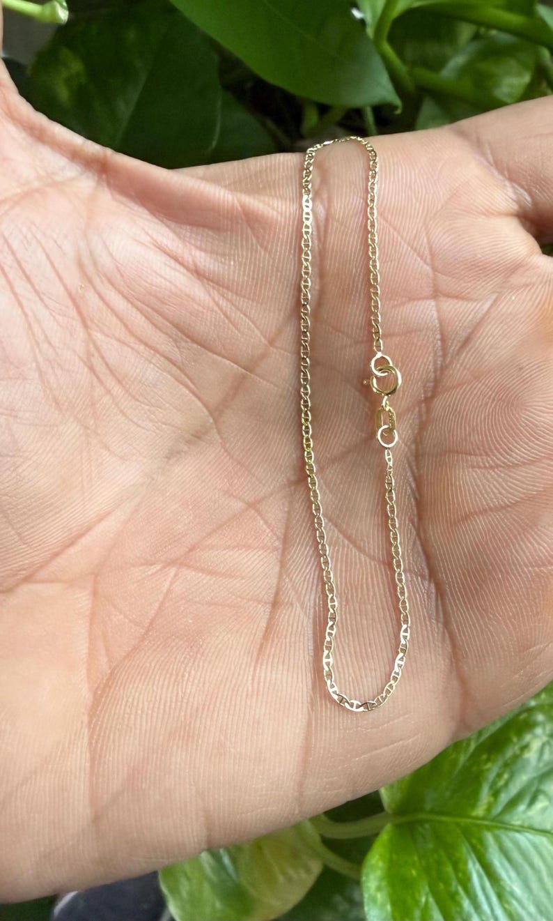 May include: A delicate gold chain bracelet featuring small, oval-shaped mariner links. The bracelet is secured with a circular spring ring clasp and includes a small rectangular metal tag near the closure, showcasing a polished metallic finish.