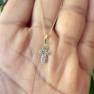 May include: A gold-colored pendant necklace featuring a small, ornate cross. The cross has intricate detailing and is suspended from a delicate chain. The necklace is displayed against a neutral background.