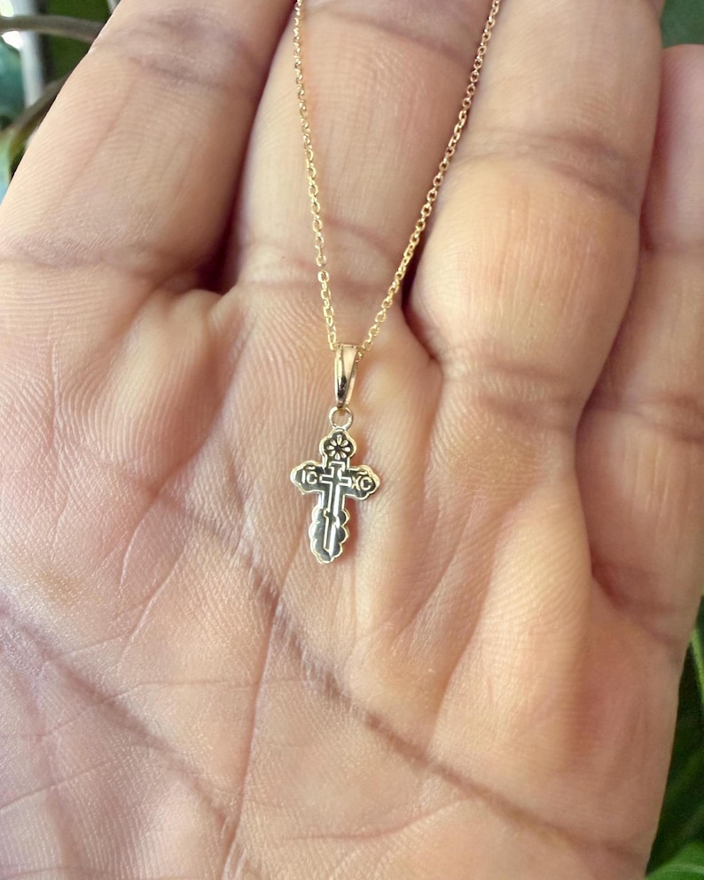 May include: A gold-colored pendant necklace featuring a small, ornate cross. The cross has intricate detailing and is suspended from a delicate chain. The necklace is displayed against a neutral background.