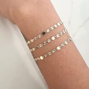 May include: Three delicate gold bracelets are displayed on a wrist. Each bracelet features a repeating pattern of small, flat, oval-shaped links interspersed with starburst-like details. The bracelets are layered, creating a subtle, elegant look.