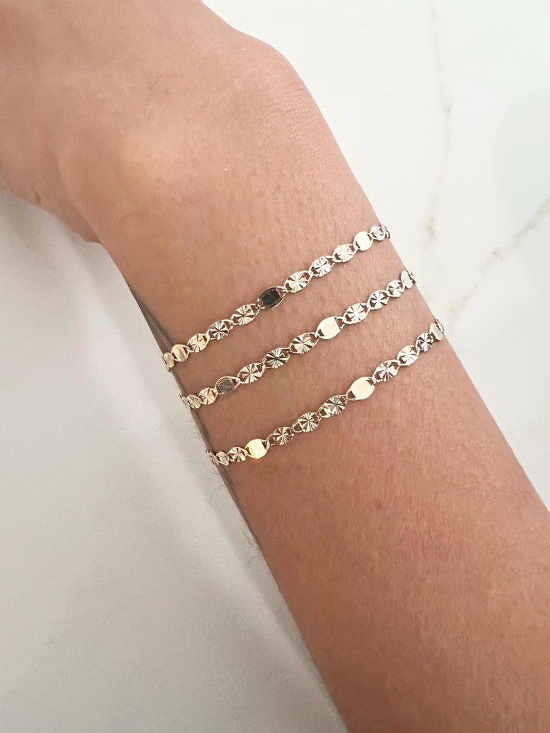 May include: Three delicate gold bracelets are displayed on a wrist. Each bracelet features a repeating pattern of small, flat, oval-shaped links interspersed with starburst-like details. The bracelets are layered, creating a subtle, elegant look.