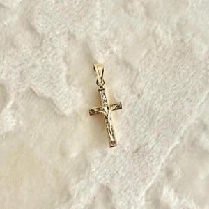Op de afbeelding: Een goudkleurige hanger in de vorm van een kruis, met een afbeelding van Jezus Christus. Het kruis is versierd met kleine, heldere edelstenen en heeft een lus aan de bovenkant voor een ketting.