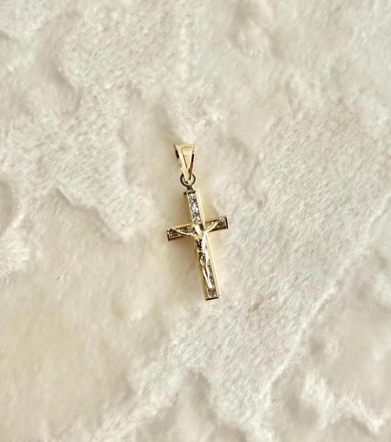 Op de afbeelding: Een goudkleurige hanger in de vorm van een kruis, met een afbeelding van Jezus Christus. Het kruis is versierd met kleine, heldere edelstenen en heeft een lus aan de bovenkant voor een ketting.