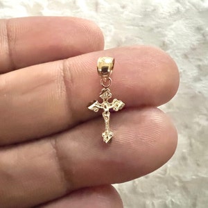 May include: A small, gold-coloured cross pendant with intricate detailing. The cross features a figure of Jesus and ornate embellishments. The pendant is suspended from a small loop. The overall design is delicate and religious.