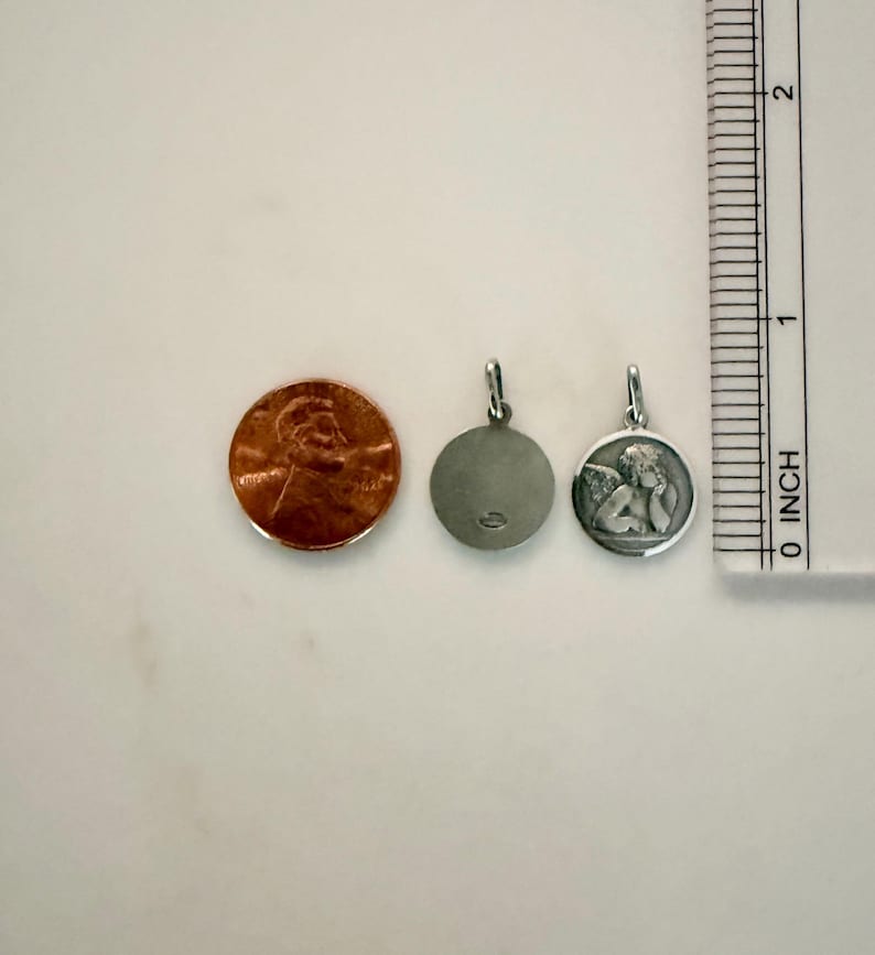 Peut inclure: Deux petits pendentifs en argent, dont un avec un ange, &agrave; c&ocirc;t&eacute; d'un penny et d'une r&egrave;gle. Le pendentif ange a un relief d&eacute;taill&eacute; d'un ch&eacute;rubin. L'autre pendentif est uni. La r&egrave;gle montre des mesures en centim&egrave;tres.