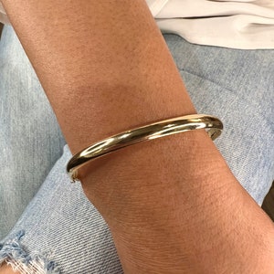 May include: A gold-colored bangle bracelet. The bracelet is smooth and rounded, with a simple, classic design. The bracelet is worn on a wrist, against a background of denim jeans and a white top.