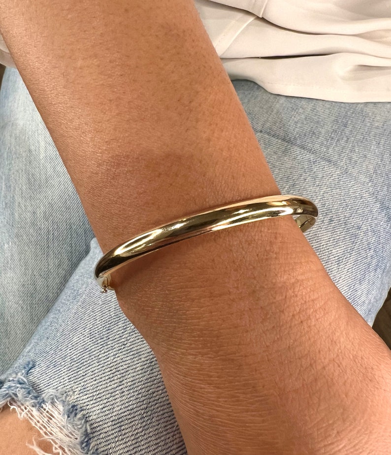 May include: A gold-colored bangle bracelet. The bracelet is smooth and rounded, with a simple, classic design. The bracelet is worn on a wrist, against a background of denim jeans and a white top.