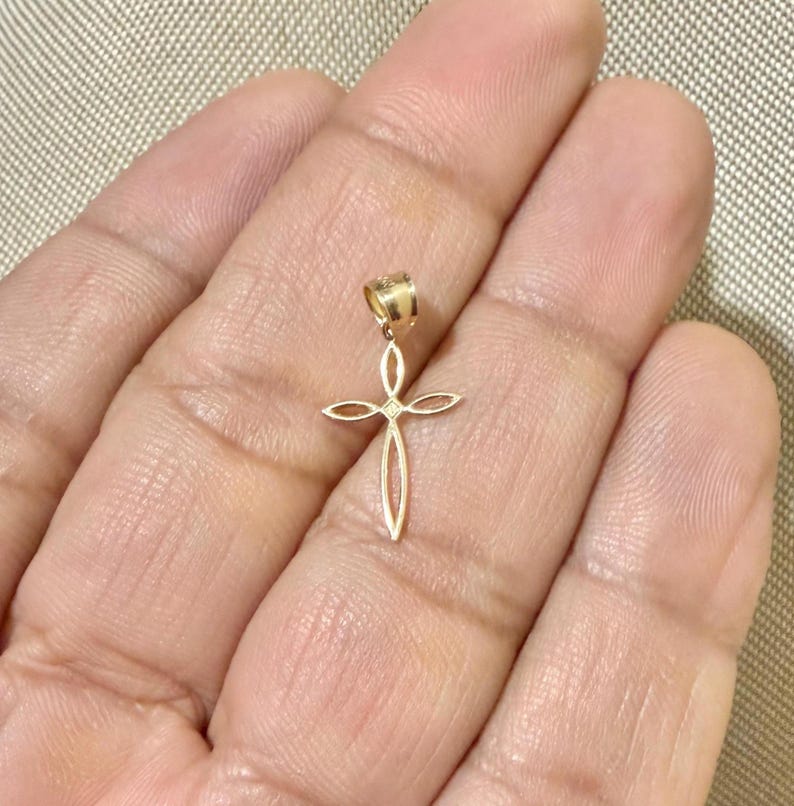Puede incluir: Un colgante dorado con forma de cruz. La cruz tiene un dise&ntilde;o calado con un peque&ntilde;o detalle en forma de diamante en el centro. El colgante cuelga de un peque&ntilde;o lazo.
