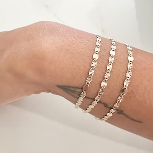 May include: Three delicate gold bracelets, each featuring a repeating pattern of oval and star-shaped links. The bracelets are displayed on a person's arm, showcasing their intricate design and reflective surface. The jewelry is set against a neutral background.